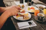 Hände greifen nach Essen auf einem weißen Teller, daneben Getränke und weitere Speisen.