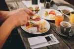 Hände greifen nach einem Teller mit Frühstück und verschiedenen Getränken auf einem Holztisch