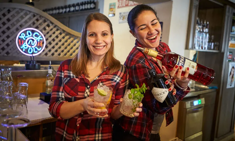 Zwei Barkeeperinnen mit Cocktails hinter der Bar, lächeln und halten Getränke in der Hand