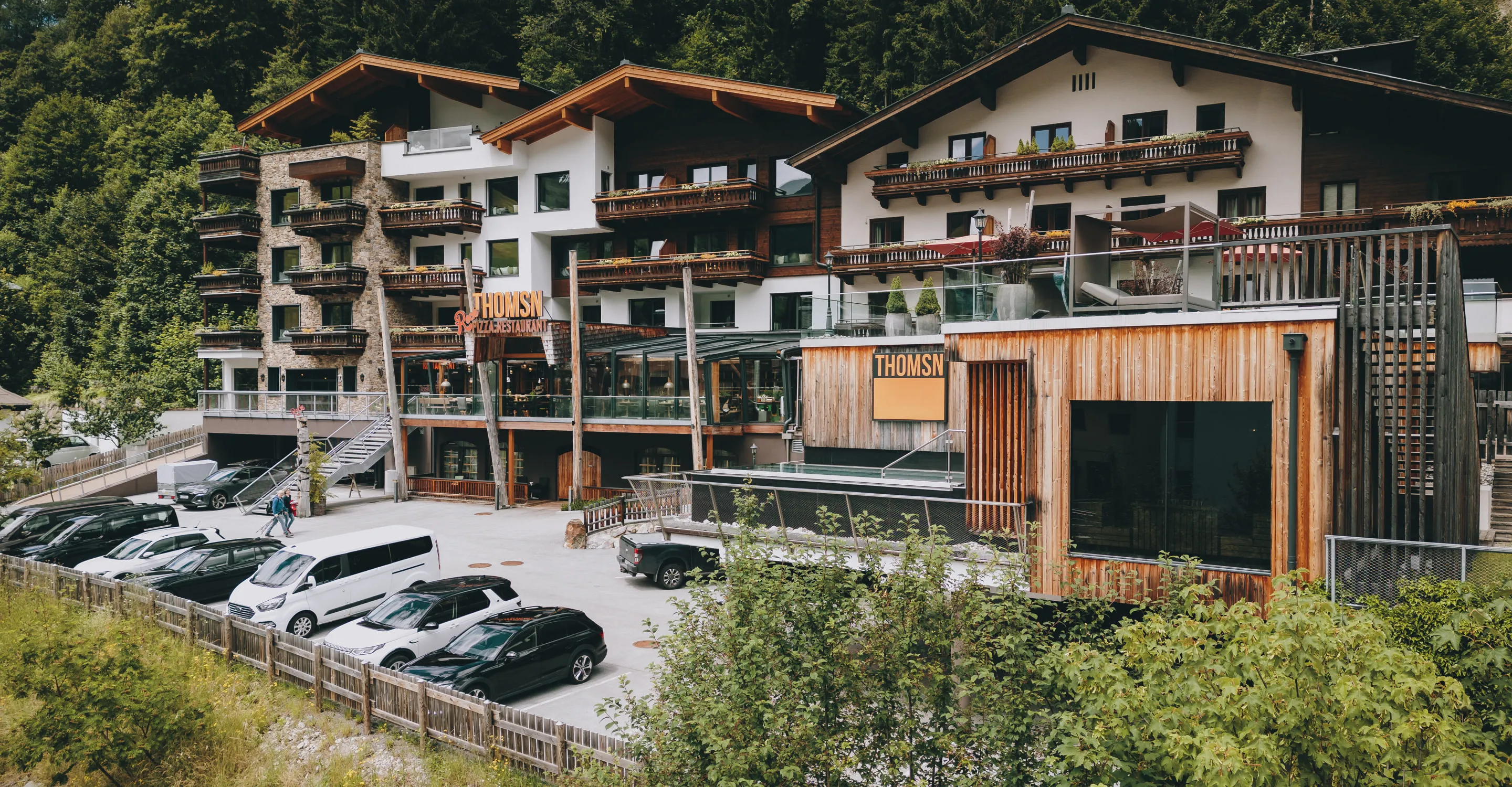 Hotel Thomsen mit Terrasse und Parkplätzen vor der Fassade, umgeben von Bäumen.