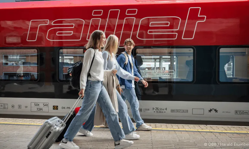 Drei Reisende gehen am Bahnhof vorbei, Zug im Hintergrund.